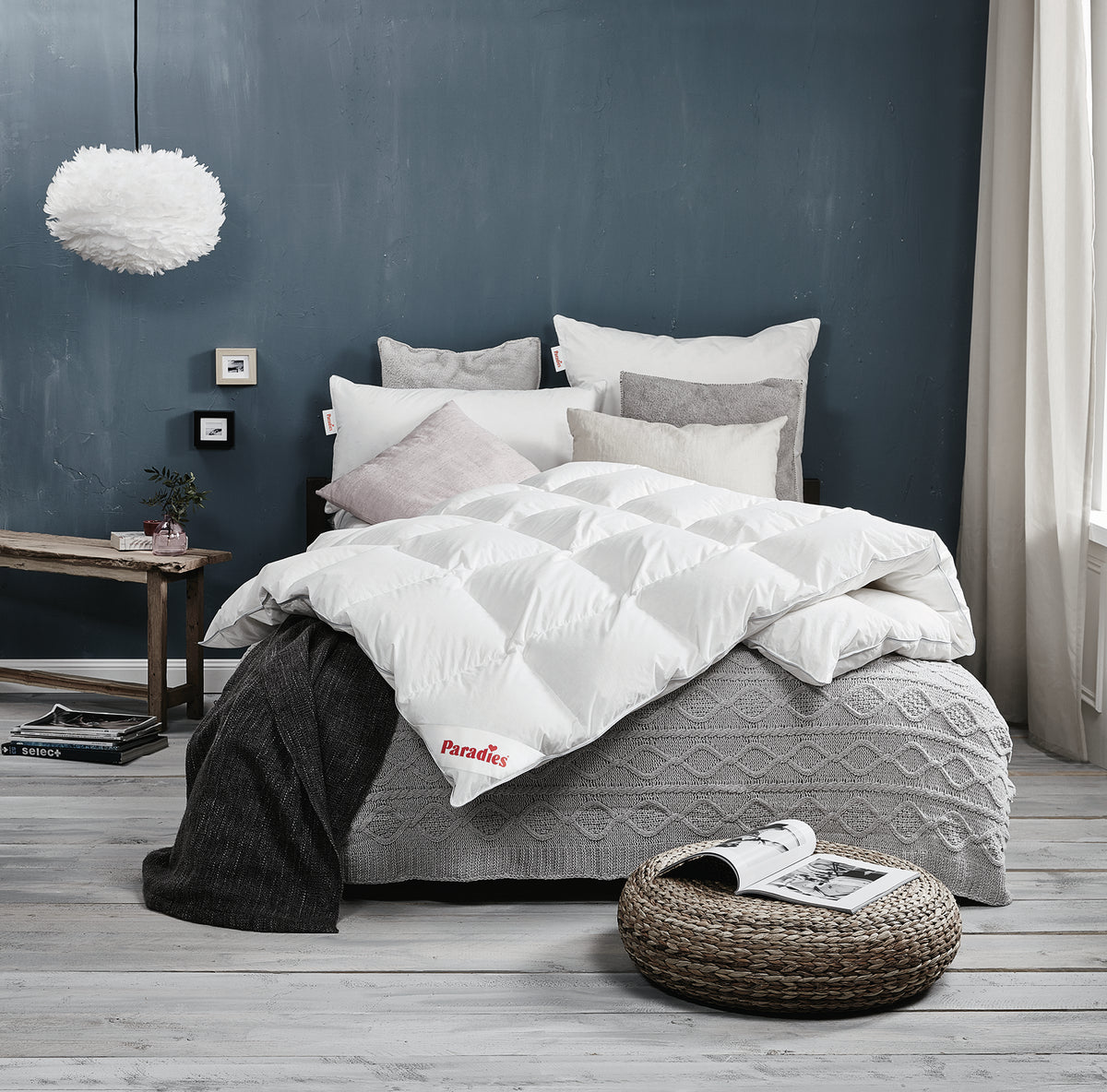Bett mit mehreren Kissen und einer weißen Steppdecke mit rotem Paradies-Logo auf einer grauen Strickdecke vor dunkelgrauer Wand